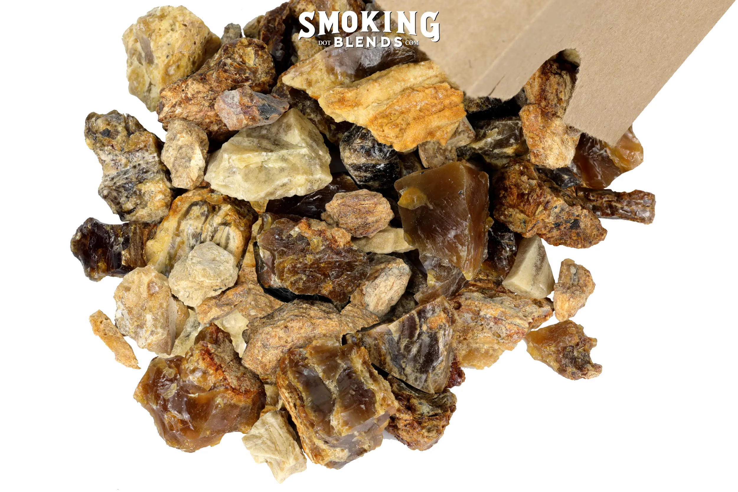 Close-up side view image of amber resin incense chunks of mixed colors, including deep mahogany, golden amber, sandy tan, and pale yellow pouring from a tin tie kraft paper bag. Pieces range from small pebbles to around an inch and a half in diameter; some are smooth and glassy, others rough and matte. At the top of the image, there is a white watermark that says Smoking Blends dot com.