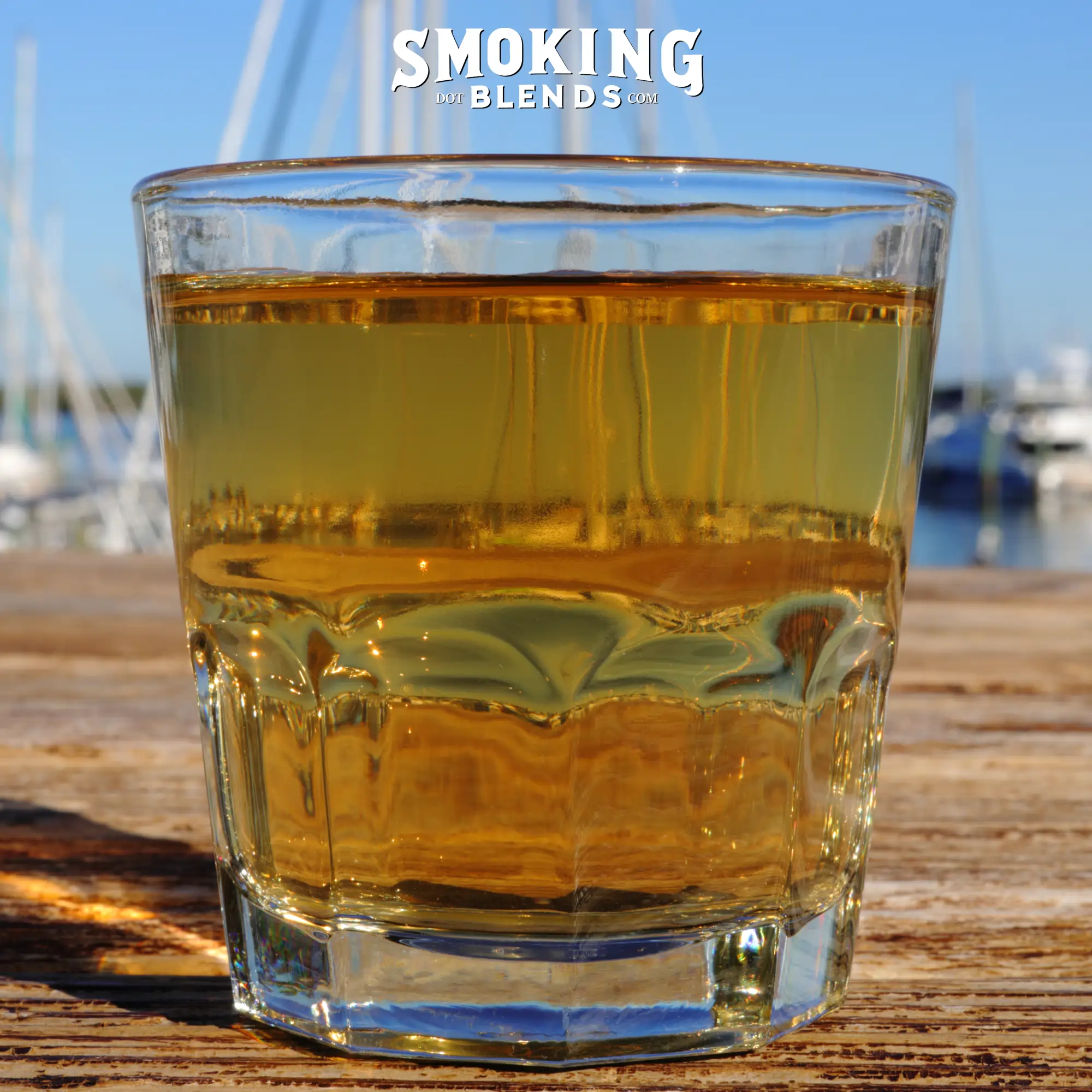 A close-up shot of a clear glass filled with brewed Klip Dagga tea, showing the golden liquid transparency against a blurred background of boats and blue sky.