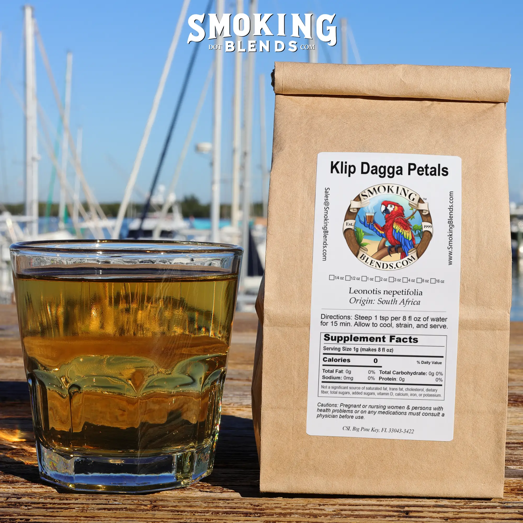 A eco friendly kraft paper package of Klip Dagga Petals standing upright next to a glass of prepared herbal tea on a sunny outdoor wooden deck at a marina.
