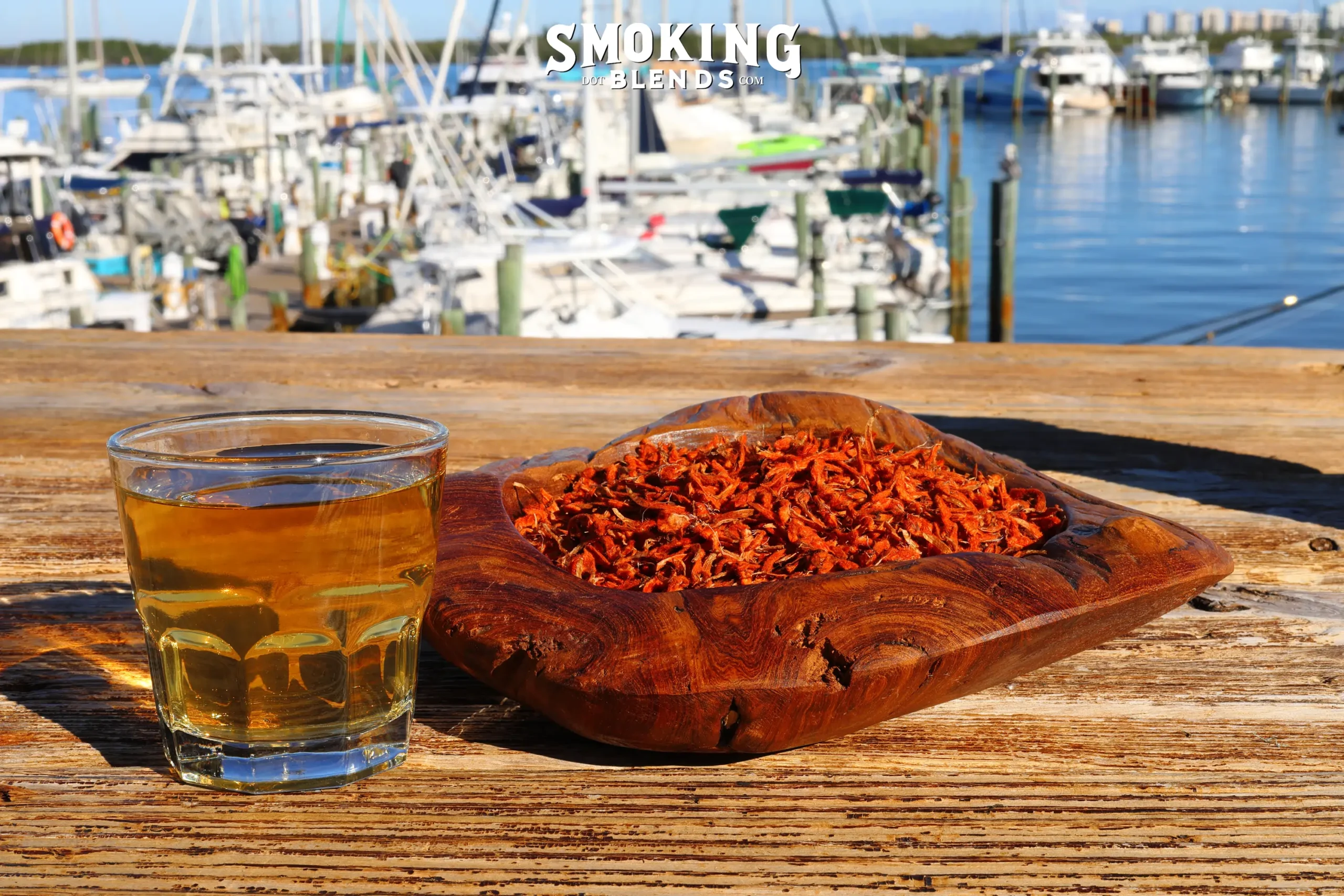A glass of golden-amber Klip Dagga tea sitting next to a carved wooden bowl filled with dried red Leonotis nepetifolia petals, set on a wooden table overlooking a sunny marina.