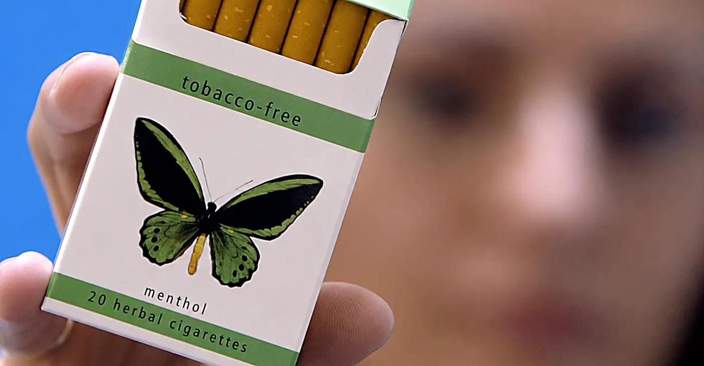 This featured image for the article "The Problem with Herbal Cigarettes and the Solution" shows a close-up of a hand holding a white pack of cigarettes. The box has green stripes at the top and bottom with the words "tobacco-free," "menthol," and "20 herbal cigarettes" printed on it, along with a drawing of a green and black butterfly right in the middle. The top of the pack is open, showing the tan filters of the cigarettes inside. In the blurry background, you can see a person's face against a bright blue background.